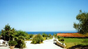 A pathway leading to a building with a red-tiled roof, surrounded by greenery and trees, overlooking a body of water. - Hotel Pashos (Kriopigi)