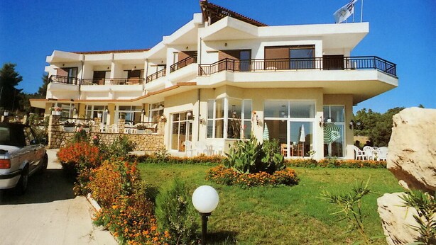 A multi-story hotel with balconies, outdoor seating, and a flag on the roof. - Hotel Pashos (Kriopigi)