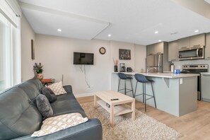 A modern living room with a black leather sofa, a wooden coffee table, and a kitchen area with a microwave, oven, and bar stools.
