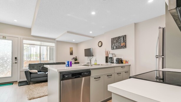 A modern kitchen with stainless steel appliances, a dishwasher, and a large island. There is a black sofa and a window with a view of the outdoors.