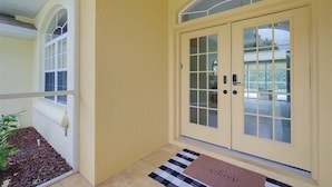 Une façade jaune avec une porte à deux battants, un paillasson et un petit jardin agrémenté d’une plante.