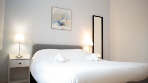 A neatly made bed with white linens and two towels, a bedside table with a lamp, and a framed artwork on the wall.