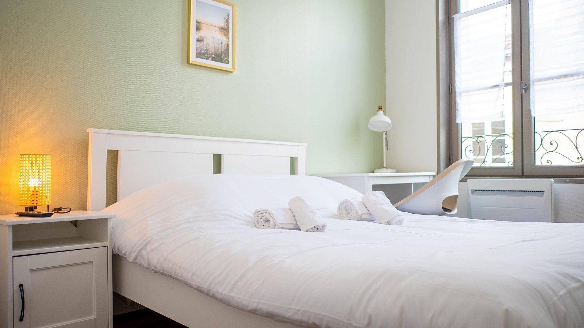 A neatly made bed with white linens and rolled towels, a bedside lamp, a small table with a lamp, and a window with a view of the outside.
