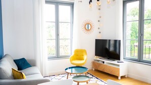 A modern living room with a sofa, a yellow armchair, a TV on a wooden stand, and large windows with a view of greenery.