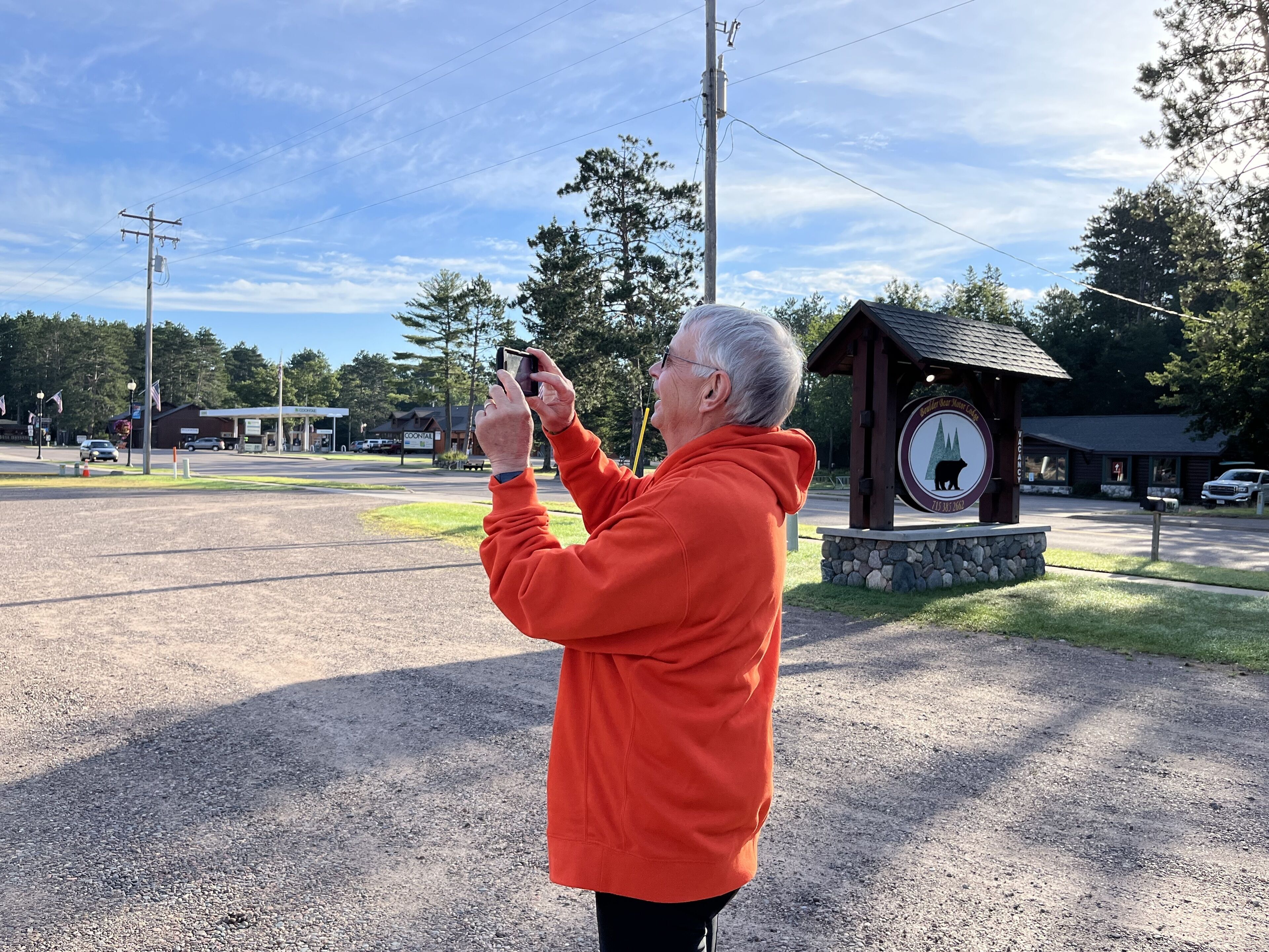 Una persona fotografa un orologio a torre in legno con l'emblema di un orso, circondato da un'area ghiaiosa con una strada e degli edifici sullo sfondo.