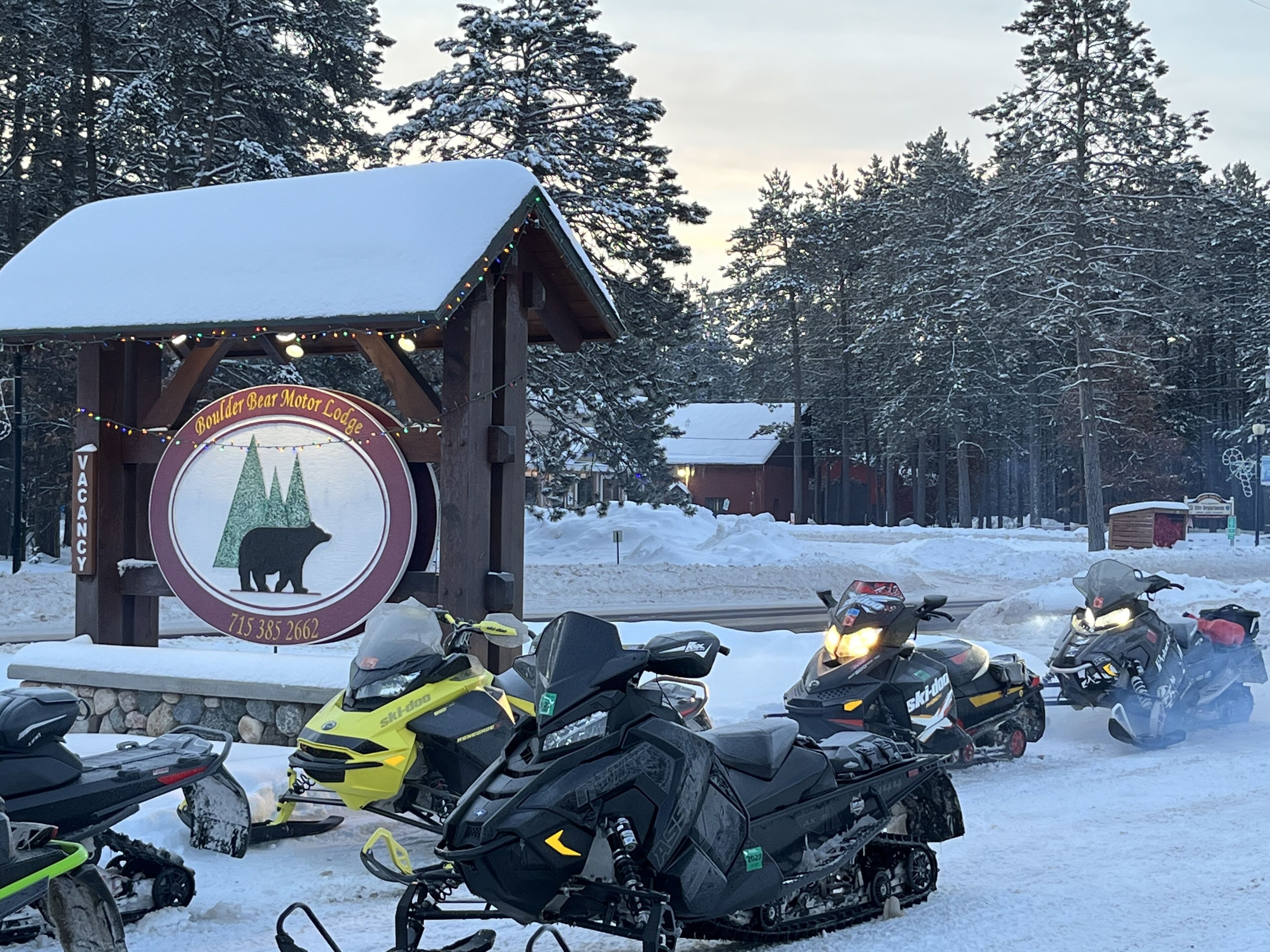 A lodge sign with a bear logo and a phone number. Snowmobiles are parked in front.