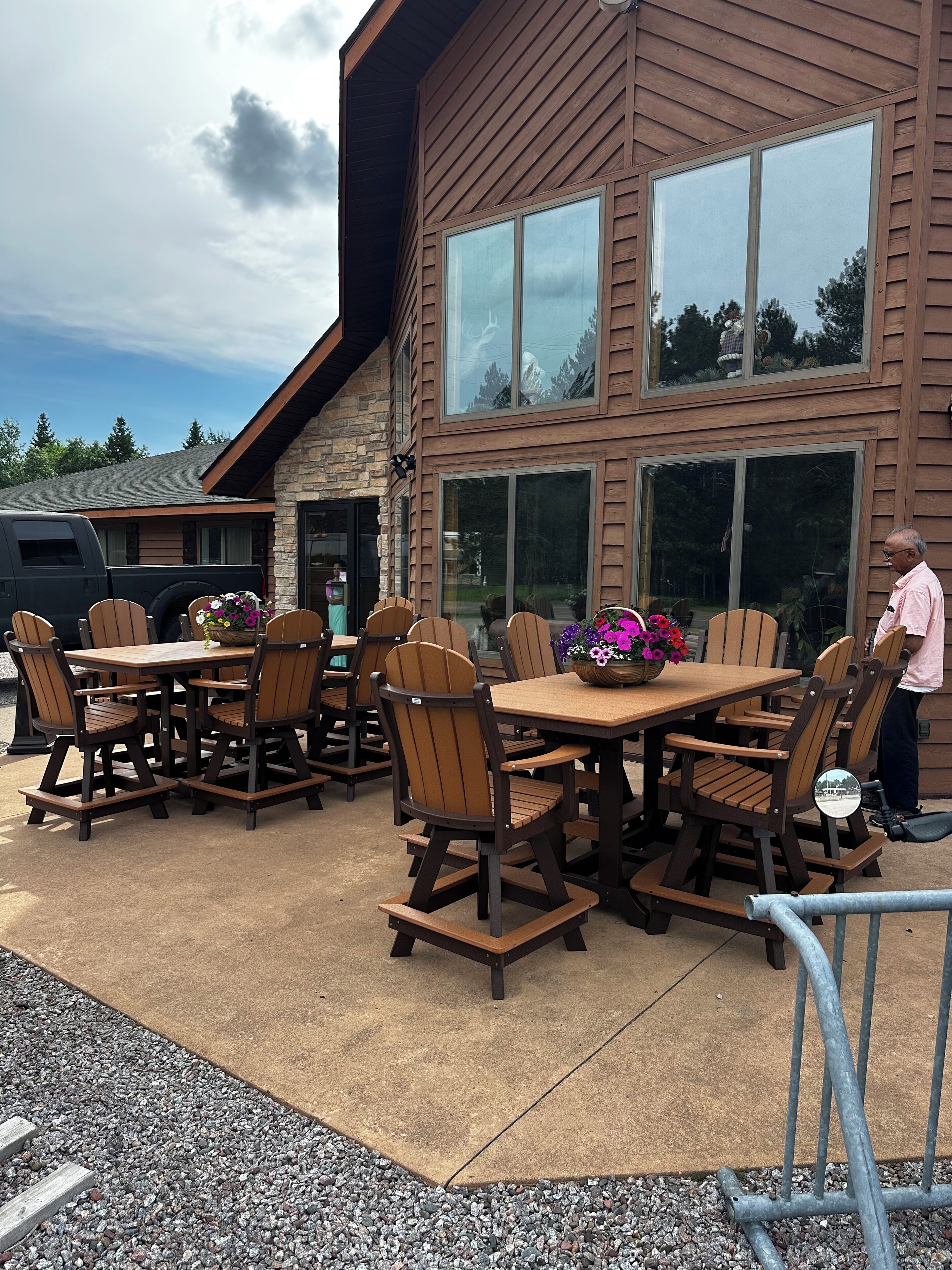 An outdoor patio with wooden tables and chairs, a stone wall, and a large window overlooking a forested area.