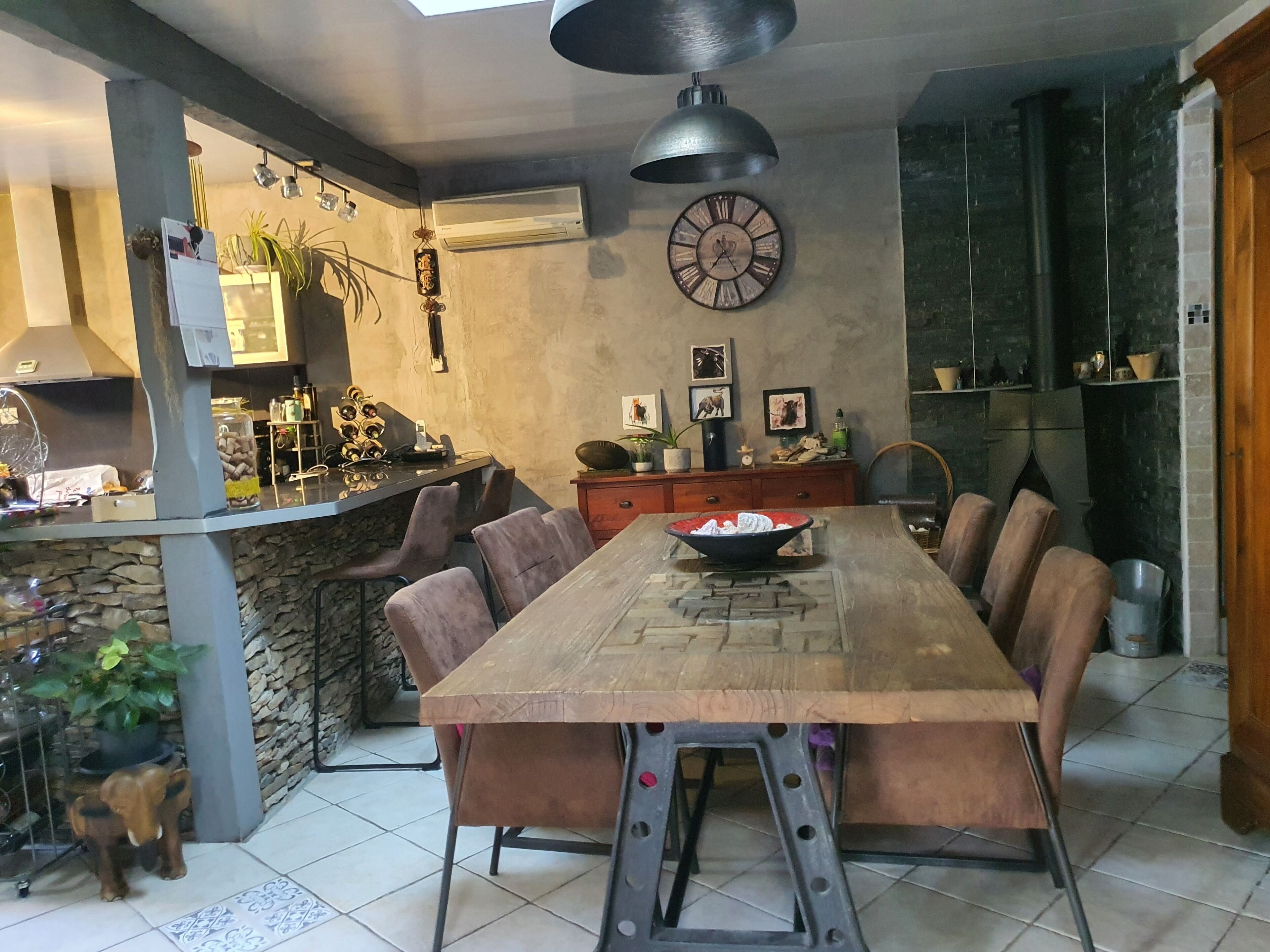 A dining area with a long wooden table, multiple chairs, a clock, and a stone wall.
