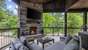 An outdoor living area with a stone fireplace, a large flat-screen TV, and a seating arrangement. - Romantic Cabin, Hot Tub, Fire Pit, Pets, Blackwater (Broken Bow)