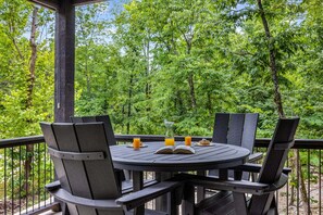 A round wooden table with four chairs, an open book, and two glasses of orange juice on a balcony with a view of dense green trees.