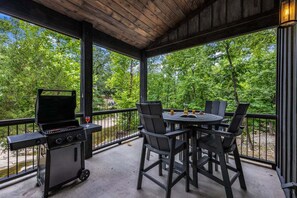 An outdoor patio with a grill, dining table, and chairs, surrounded by a wooden deck and a view of trees.