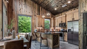 A rustic kitchen with wooden walls, a central island with bar stools, a large refrigerator, and a view of greenery outside. - Romantic Cabin, Hot Tub, Fire Pit, Pets, Blackwater (Broken Bow)