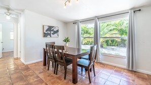 Ein Essbereich mit Holztisch und Stühlen, einem Deckenventilator, einer an der Wand montierten Lampe und einem großen Fenster mit Vorhängen.
