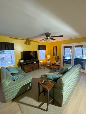 A living room with a sofa set, a television, a ceiling fan, and a window with blinds.
