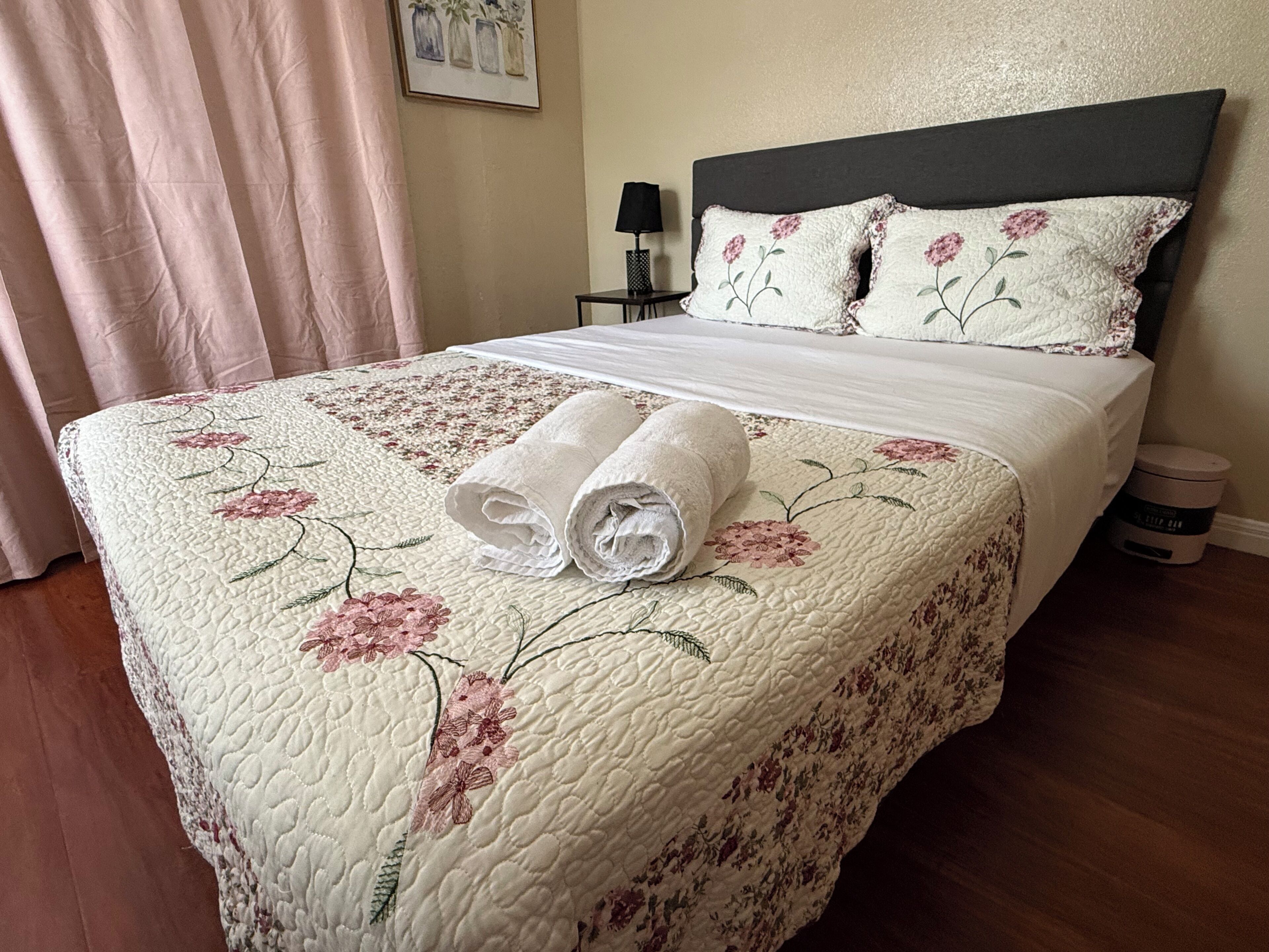 A neatly made bed with a floral quilt, a pair of rolled towels, a bedside lamp, and a small table with a lamp.