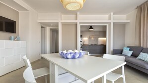 Un salon moderne avec une table à manger blanche, des chaises blanches, un canapé gris agrémenté de coussins à motifs, et une cuisine en arrière-plan.