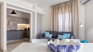 Un salon moderne avec un canapé gris, des coussins à motifs bleus, une table blanche sur laquelle se trouve un bol en céramique, et une cuisine en arrière-plan.