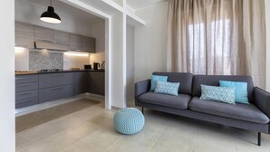 Un salon moderne avec un canapé gris, deux coussins à motifs, un pouf rond et une grande fenêtre agrémentée de rideaux beige.