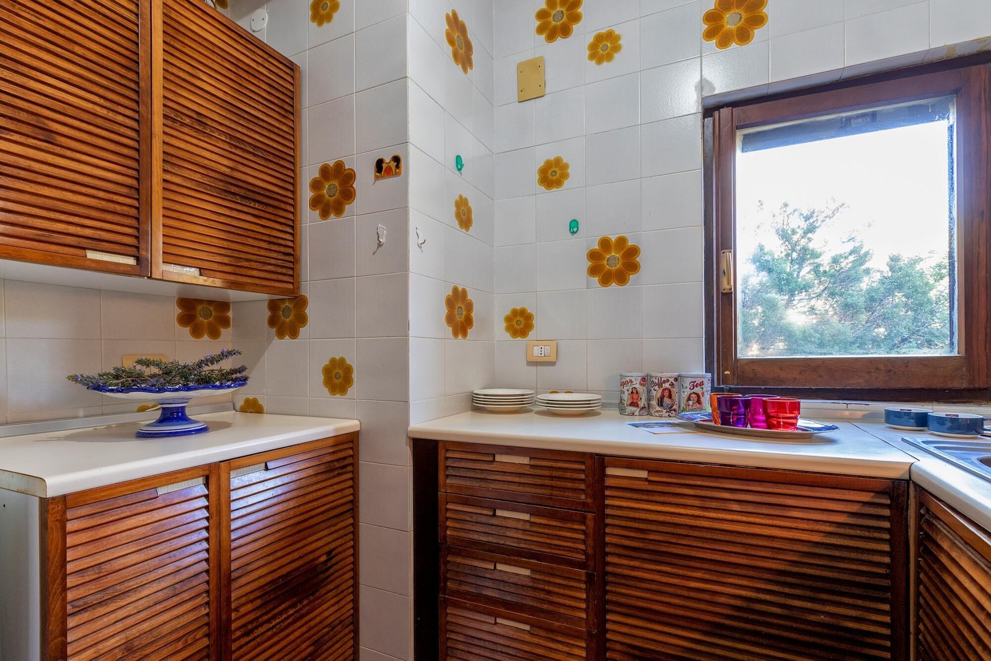 Una cucina con mobili in legno, piastrelle bianche decorate con motivi floreali e una finestra vista alberi.