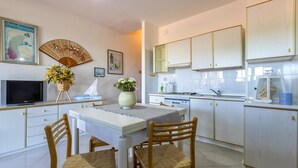 A modern kitchen with white cabinets, a dining table with chairs, a TV, and decorative items like a fan and flowers. - Ss-m290-cdva0at - Villino Punta Negra (Stintino)