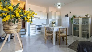 A modern kitchen with white cabinets, a dining table, and chairs. There is a wicker basket with yellow flowers on a stand. - Ss-m290-cdva0at - Villino Punta Negra (Stintino)