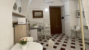 A compact kitchen with a microwave, sink, and a small refrigerator. There is a dining table with a vase of flowers and a chair. The floor has a checkered pattern.