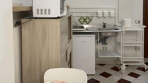 A compact kitchenette with a microwave, a small refrigerator, and a sink. There is a chair with a table in front, and a decorative item on the wall.