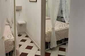 A narrow hallway with a checkered floor, a bed with a floral bedspread, and a small shelf with a lamp. - Sr-f943-cavo83at - La Casa di Benso (Noto)
