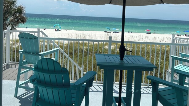 Un balcon avec des chaises turquoise et une table, donnant sur une plage parsemée de parasols et animée par la présence de nombreuses personnes.