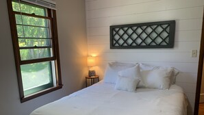 Een slaapkamer met een groot bed, een raam met uitzicht op bomen, een decoratief object aan de muur en een klein nachtkastje met een lamp.