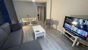 A modern living room with a grey sofa, a white coffee table, a TV on a stand, and a dining area with a table and chairs. - Cosy, Modern Apartment (Darlaston)