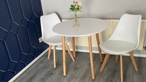A room with a round white table and two white chairs with wooden legs. There is a vase with flowers on the table. The room has a grey wooden floor and a dark blue wall with a geometric pattern. - Cosy, Modern Apartment (Darlaston)