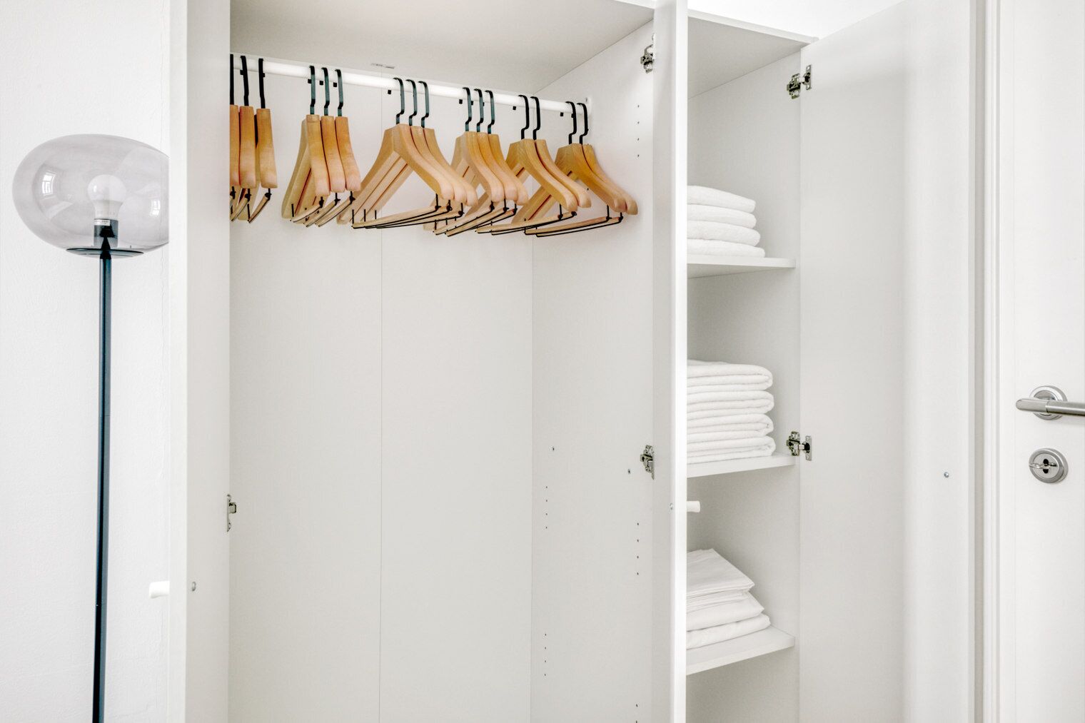 Een witte inloopkast met houten kleerhangers en netjes opgevouwen witte handdoeken op planken.