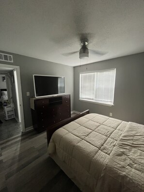 Relax in this serene bedroom with a large bed, dresser, and TV, featuring a window with blinds and a ceiling fan.