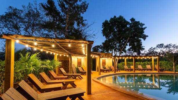 Un área junto a la piscina con tumbonas de madera y una pérgola con luces colgantes.
