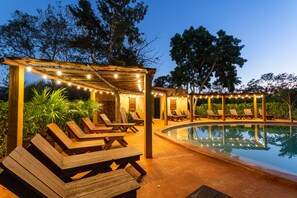 Un área junto a la piscina con tumbonas de madera y una pérgola con luces colgantes.
