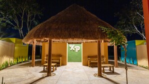Estructura con techo de paja, puerta verde y letras 'XP', rodeada de bancos y una zona pavimentada.
