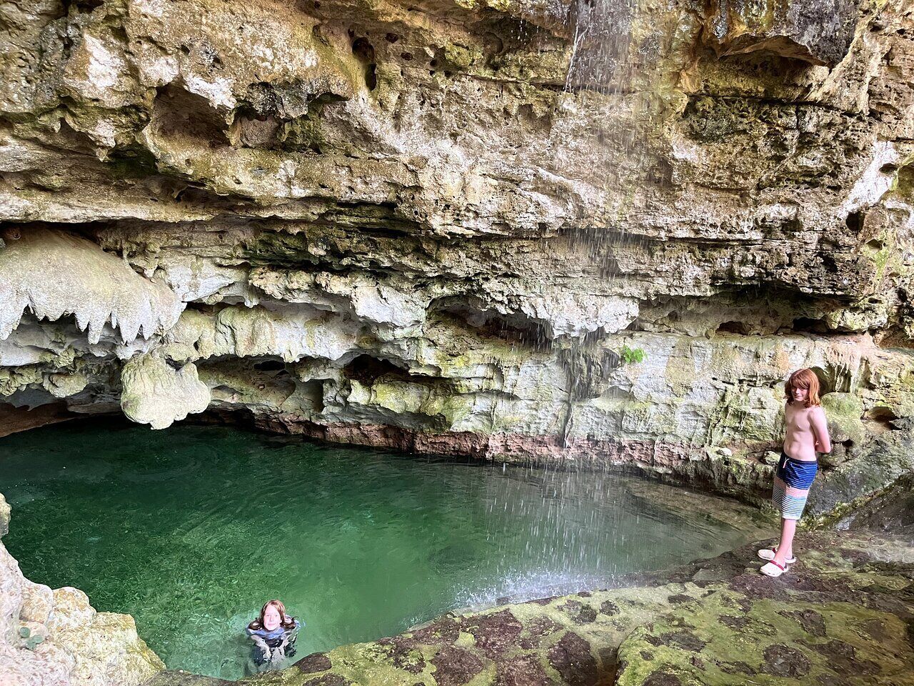 Una cueva con una poza cristalina y una persona de pie en un saliente.