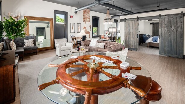 A spacious living room with a large wooden ship's wheel table, a ceiling fan, and a modern kitchen in the background.