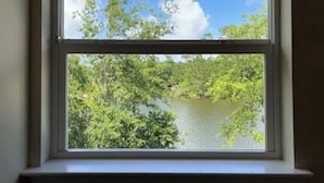 Une fenêtre donnant sur un lac, bordé d’une végétation luxuriante, et sur un ciel bleu dégagé.