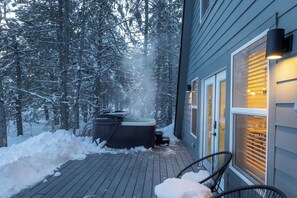 A wooden deck with a hot tub and chairs, surrounded by snow-covered trees.