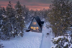 A cabin with a sloped roof, surrounded by snow-covered trees, and a clear path leading to the entrance.