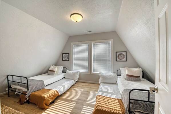 A small, well-lit attic bedroom with two single beds, a window with blinds, and a ceiling light.