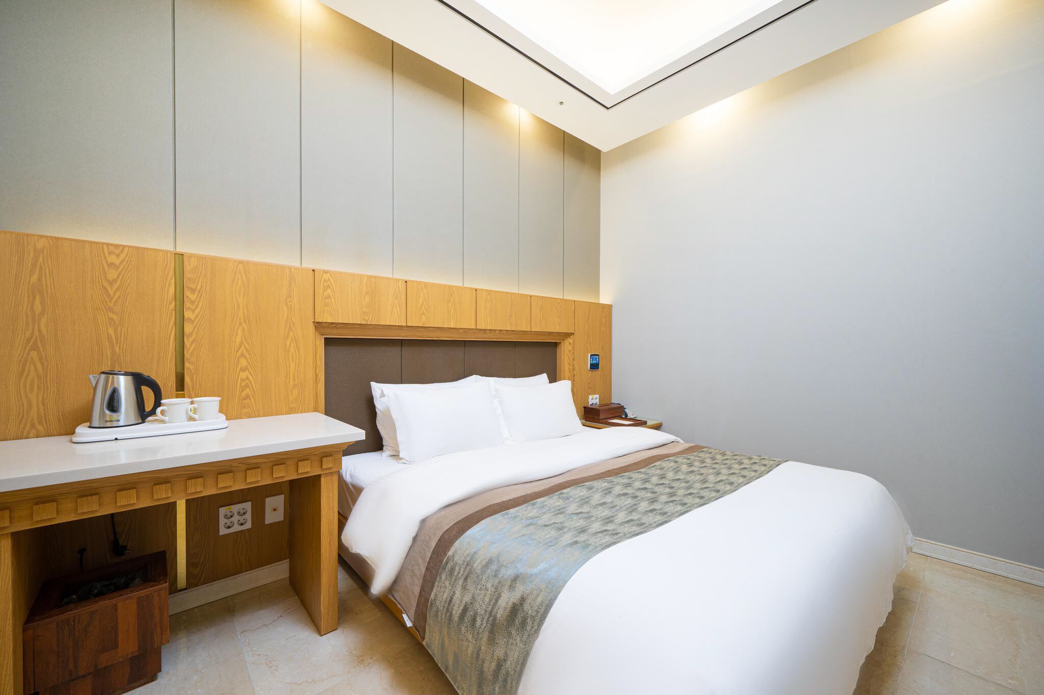 A hotel room with a bed, a wooden desk, a nightstand, and a skylight.