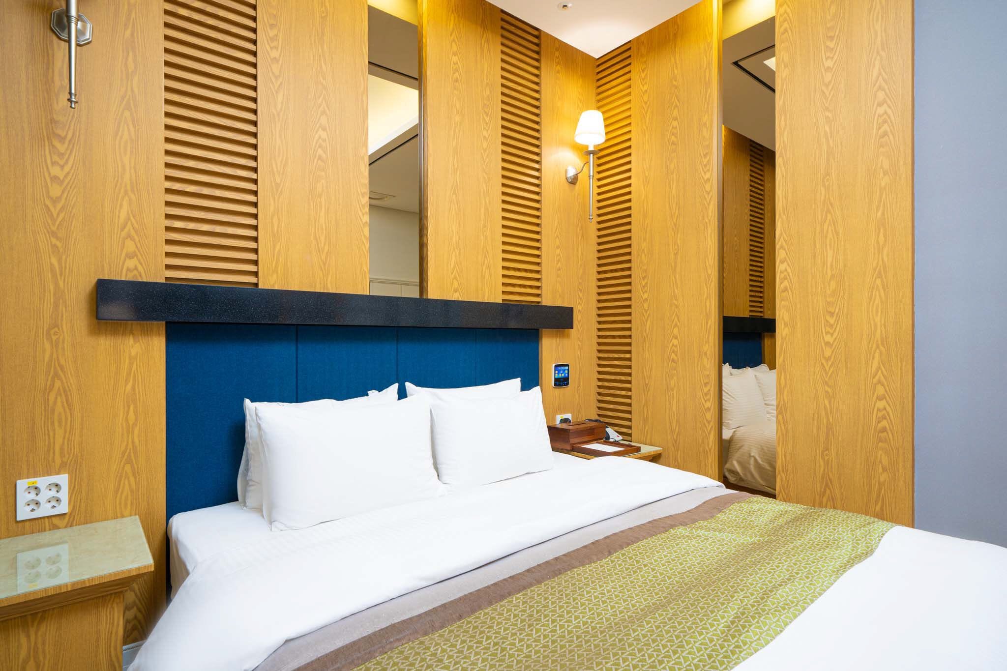 A hotel room with a wooden panel wall, a blue headboard, a white bed with a patterned throw, a bedside table with a lamp, and a mirror.