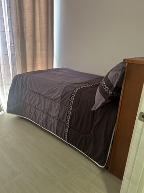 A single bed with a dark-colored bedspread and a decorative pillow. The bed is positioned against a wall with a wooden headboard.
