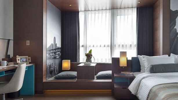 A modern hotel room with a large bed, a desk with a chair, a small seating area with a plant, and a cityscape view through the window. - Merciel Hotel (Busan)