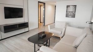 Un salon moderne avec un téléviseur à écran plat fixé au mur, une table basse noire, un canapé beige et un fauteuil blanc.