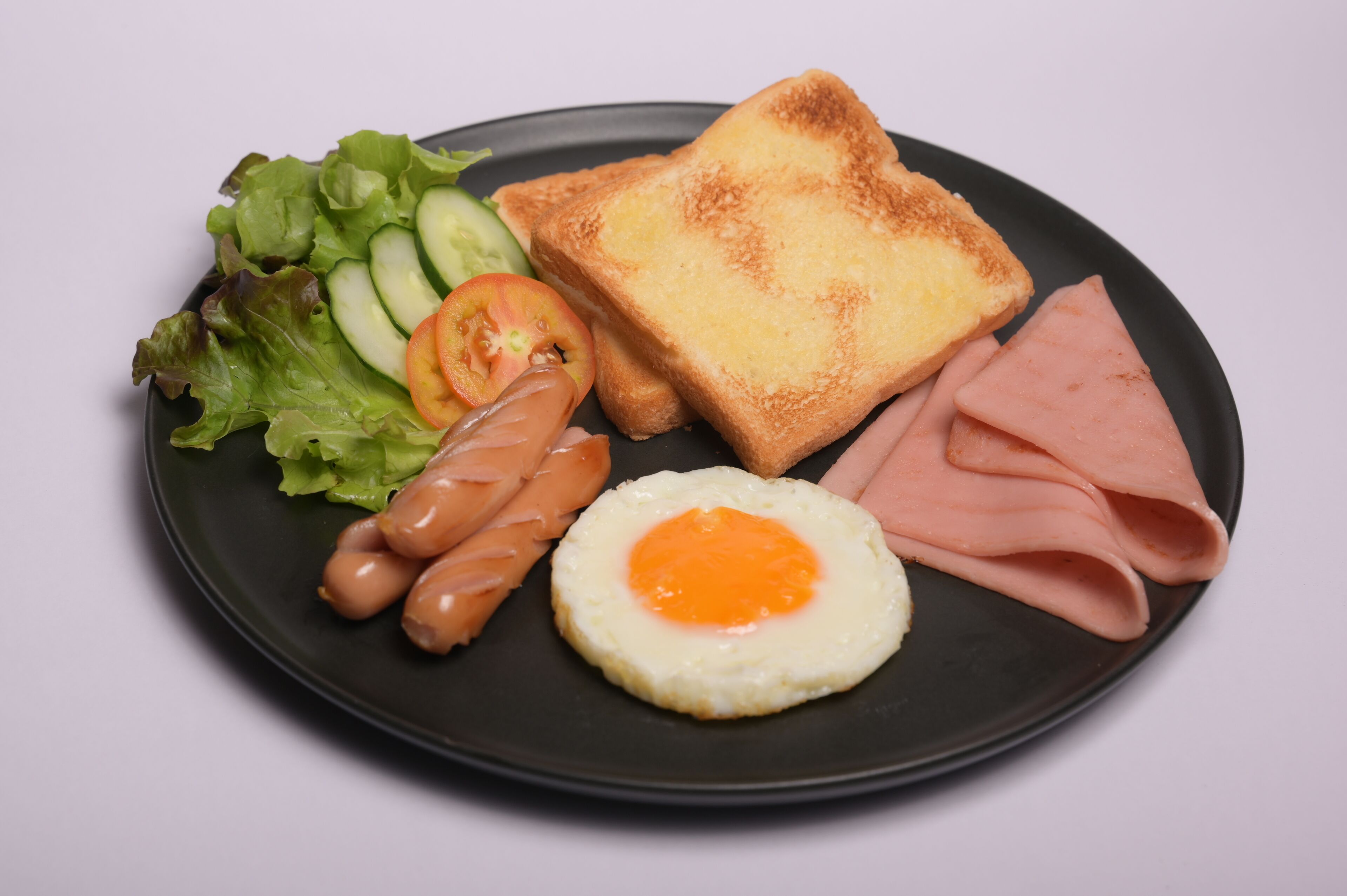 Ein Teller mit Toast, Schinken, einem Spiegelei, Würstchen, Salat und Gurkenscheiben.