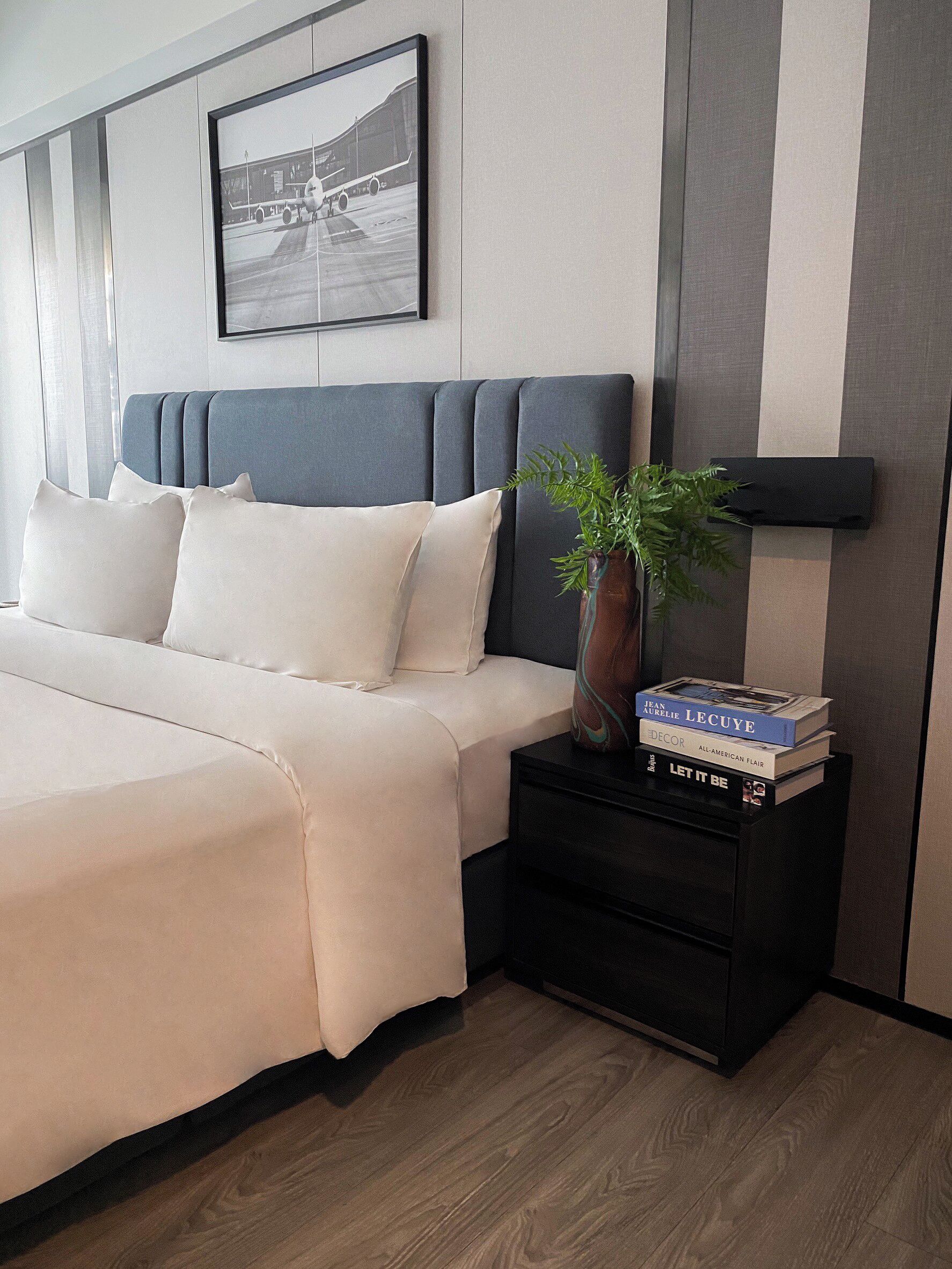 Een netjes opgemaakt bed met een grijs hoofdbord, een nachtkastje met een plant en boeken, en een ingelijste foto aan de muur.
